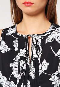 Svart blus med vitt blommönster, rund halsringning och nyckelhålsdetalj framtill. Har texturerade veck och knytstängning.