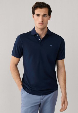 Jeune homme aux cheveux foncés portant un polo bleu marine et un pantalon bleu clair, debout devant un fond neutre.
