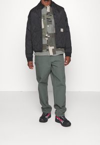 Zwarte gewatteerde jas, camouflageblouse met knopen, olijfgroene broek en zwarte sneakers met rode accenten. Focus op functionaliteit en laagjes.