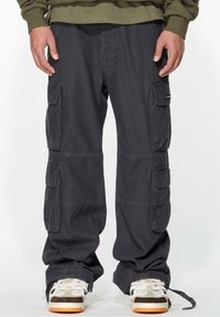 Dark cargo pants featuring multiple side pockets, a loose fit, and durable cotton material. Paired with white and brown sneakers.