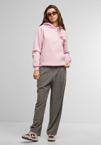Sudadera con capucha rosa y un bolsillo frontal, combinada con pantalones grises holgados y zapatillas marrones. Gráfico rojo destacado en la sudadera.