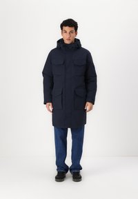 Parka marine imperméable avec capuche, dotée de plusieurs poches poitrine et latérales, d'une coupe mi-longue et d'une fermeture à boutons-pression à l'avant.