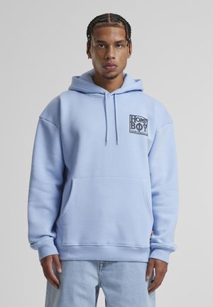 Noor mees punutud juustega, kes kannab hele sinist kapuutsiga pusa, millel on "HOME BOY loud couture" logo, ja helesiniseid teksaseid, seisab ühevärvilise taustaga.