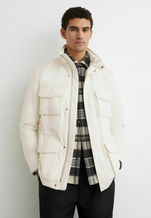 Homme portant une veste matelassée blanche par-dessus une chemise à carreaux noire et blanche, debout contre un mur clair uni, les mains dans les poches.