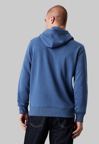 Felpa di cotone blu con un grande cappuccio, polsini e orlo a costine. Texture liscia, design semplice e vestibilità comoda. Abbinata a jeans scuri.