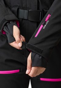 Black wetsuit with pink zipper accents and Velcro cuffs. Features a belt and a detailed sleeve opening, showcasing a smooth texture.