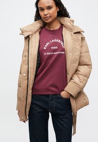 Chaqueta acolchada beige con textura acolchada, cuello alto y cierres a presión, llevada sobre una camiseta burdeos con texto gráfico blanco, combinada con jeans oscuros.