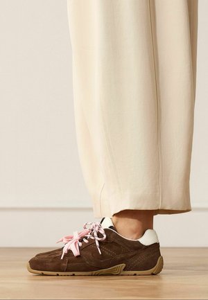 Bruine suède sneaker met witte hiel en roze veters gedragen met crèmegekleurde broek, staand op een houten vloer.