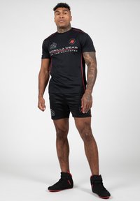 Zwarte sportshirt met rode accenten, voorzien van tekst en logo. Gepaard met zwarte shorts en zwarte sneakers met rode details. Mannelijke figuur.