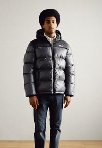 Puffy svart dunjakke med hette, med horisontale stikninger, glidelås foran og merkevarelogo. Båret over mørke jeans.