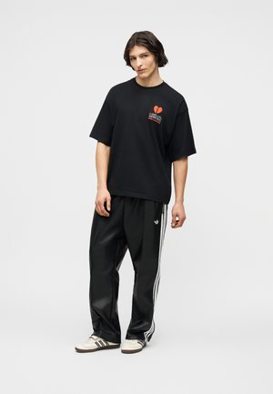 Mladić s tamnom kosom srednje duljine, nosi crnu široku majicu s grafikonom i crne Adidas hlače s bijelim prugama.