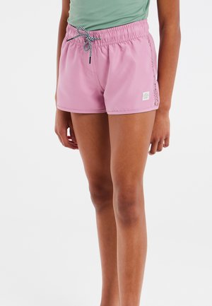 Jonge persoon draagt lichtroze korte broek met een zwart-wit trekkoord en een lichtgroen topje tegen een witte achtergrond.