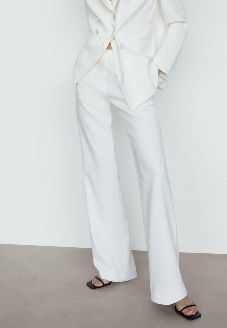Femme portant un pantalon blanc à jambes larges et un blazer assorti avec des sandales noires à talons carrés ouverts, debout sur un sol neutre.