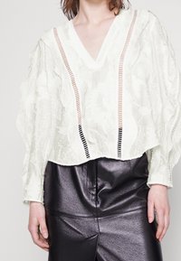 Blusa bianca a trama con scollatura a V e maniche lunghe, caratterizzata da righe verticali in pesca e nero. Abbinata a pantaloni neri lucidi.