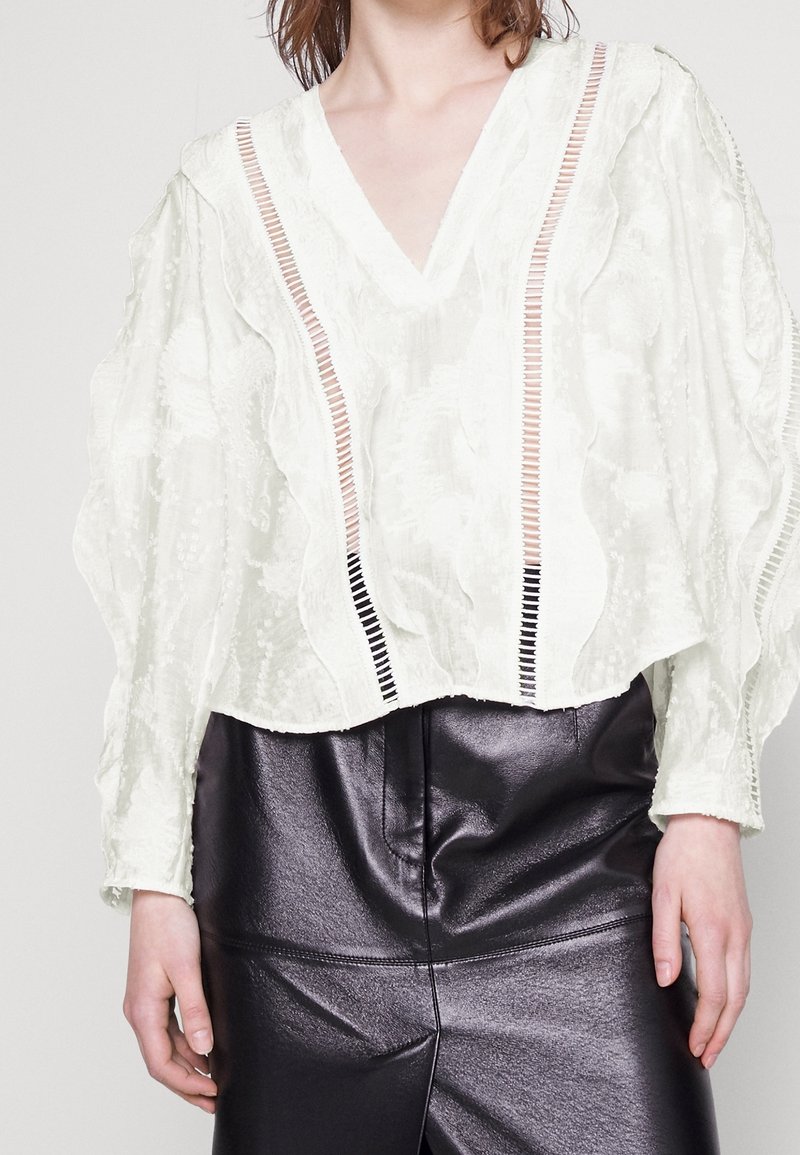 Blusa bianca a trama con scollatura a V e maniche lunghe, caratterizzata da righe verticali in pesca e nero. Abbinata a pantaloni neri lucidi.