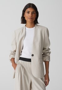 Beige blazer met een relaxed silhouet, sluiting met een enkele zwarte knoop en opgerolde mouwen, gecombineerd met een witte T-shirt en bijpassende broek.