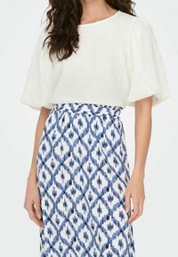 Blouse blanche texturée à manches bouffantes associée à une jupe bleue et blanche à motifs comprenant des formes losanges et des rayures horizontales.