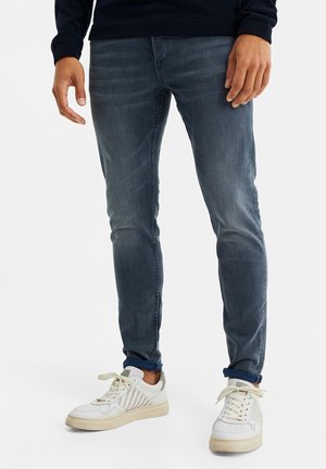 Man draagt strakke, vervaagde blauwe jeans en witte veterschoenen, staand tegen een eenvoudige lichte achtergrond.