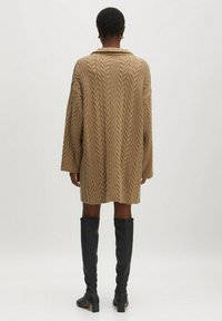Beige Strickmantel mit einem übergroßen Kragen, langen Ärmeln und strukturiertem Muster. Getragen über kniehohen schwarzen Lederstiefeln.