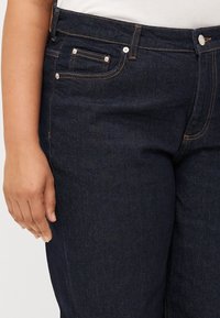 Close-up of a hand resting on dark blue jeans, featuring a subtle light stitching and front pocket details.