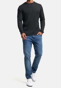 Mörkgrå kabelstickad tröja med långa ärmar, i kombination med ljusblå jeans och vita sneakers. Modell står framför en neutral bakgrund.