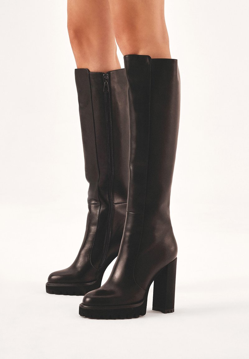 Bottes en cuir noir arrivant au genou, avec un talon carré, une fermeture éclair sur le côté et une semelle en caoutchouc texturée. Surface lisse et design minimaliste.