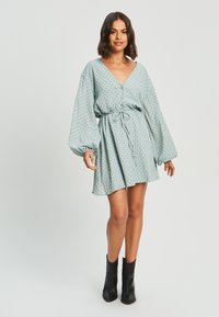 Svetlo modra obleka z V-izrezom, dolgimi balonastimi rokavi, zoženim pasom in majhnimi črnimi pikami. V kombinaciji s črnimi gležnjarji.