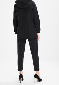 Parka negra con capucha, con la cintura ajustada, combinada con pantalones negros ceñidos y zapatos de tacón altos negros. Tejido suave, diseño minimalista.