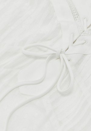 Zbliżenie na biały materiał z subtelnymi pionowymi paskami, związaną koronkową kokardą przewleczoną przez przelotki oraz ozdobnymi detalami przeszyć.