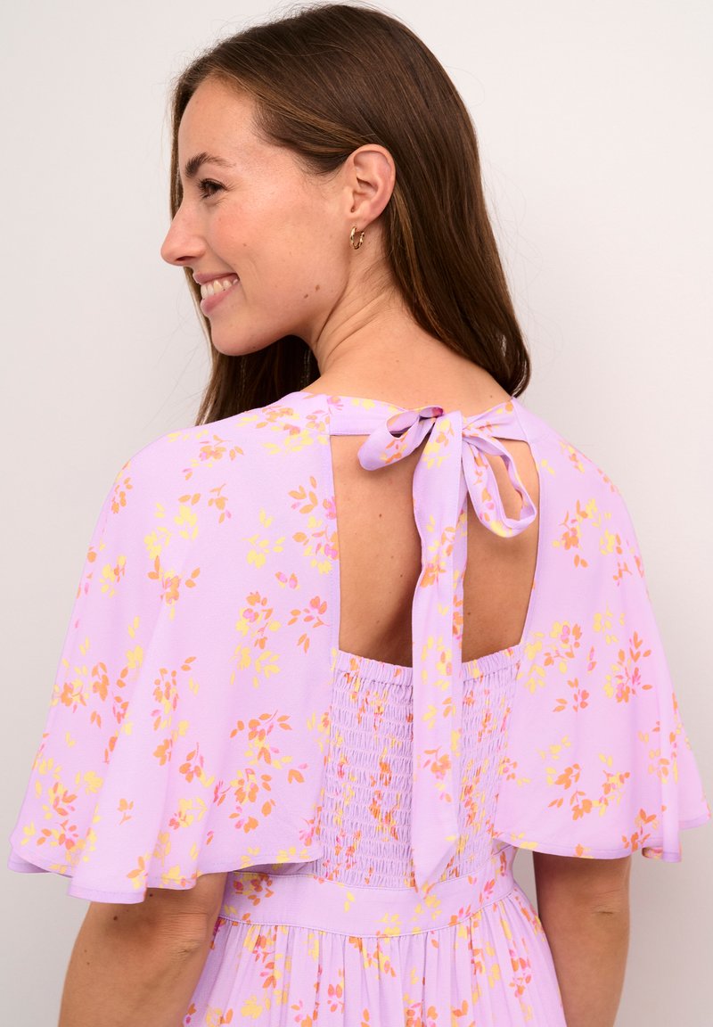 Robe lilas avec des motifs floraux ; présente des manches flottantes, un dos nu avec un lien, et un corsage froncé pour une silhouette fluide.