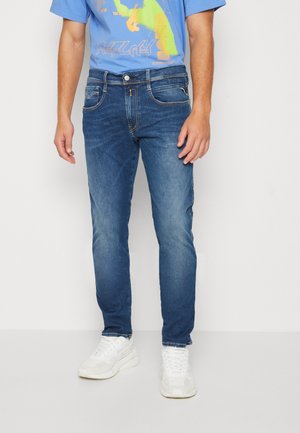 Jeans de mezclilla azul con corte ajustado, ligeras decoloraciones, cinco bolsillos y costuras blancas. Llevados con una camiseta gráfica azul y zapatillas blancas.