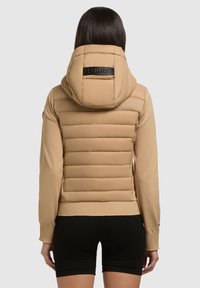 Beige pufferjakke med en figursyet silhuet, quiltet tekstur og hætte. Har et logoaccent på ryggen og ribbede manchetter.