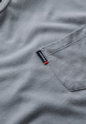 T-shirt grigio in cotone con taschino sul petto, texture morbida e una piccola etichetta nera con dettagli rossi marchiata "SUPERDRY."