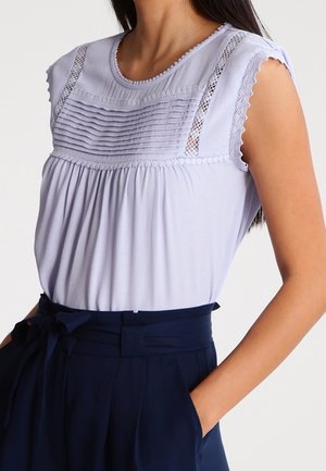 Blouse lavande sans manches avec détails plissés et en dentelle, associée à un pantalon bleu marine taille haute avec ceinture nouée.