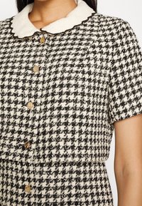 Femme portant une robe à manches courtes en pied-de-poule noir et blanc avec des boutons dorés et un col blanc festonné. Seule la partie supérieure du corps est visible.