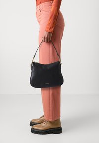 Borsa a tracolla in pelle nera con una forma morbida e arrotondata, rifinita con hardware dorato, tenuta da una persona che indossa pantaloni rosa a gamba larga e scarpe beige.