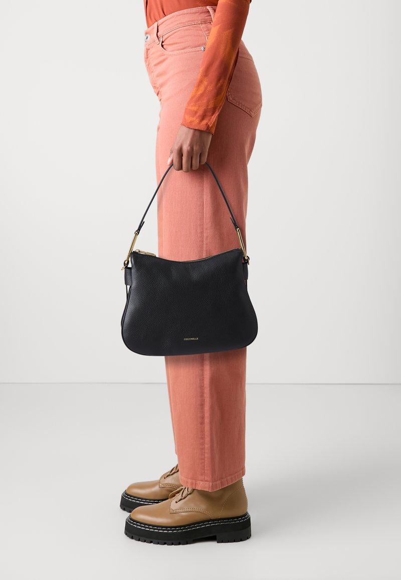 Borsa a tracolla in pelle nera con una forma morbida e arrotondata, rifinita con hardware dorato, tenuta da una persona che indossa pantaloni rosa a gamba larga e scarpe beige.