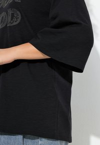 Czarna koszula z krótkim rękawem o luźnym kroju, z teksturowanym wzorem z przodu i rozcięciami po bokach. Nosi się ją na jasnoniebieskie dżinsy.