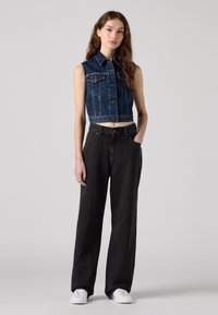 Žena koja nosi bezrukavnu tamnu traper jaknu, široke crne traperice i bijele tenisice, stoji s jednom rukom u džepu protiv obične pozadine.