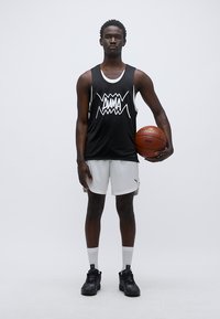 Μαύρο αμάνικο μπασκετικό φανελάκι με λευκό σχέδιο, σε συνδυασμό με λευκές βερμούδες. Κρατάει μία μπάλα του μπάσκετ, φοράει μαύρα αθλητικά παπούτσια και λευκές κάλτσες.