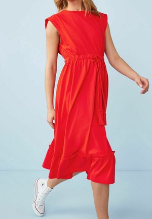Femme portant une robe midi rouge sans manches avec une taille à nouer et un ourlet froncé, accompagnée de baskets montantes blanches, sur un fond bleu clair.