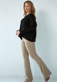 Czarny sweter z dzianiny o oversize'owym kroju, z opadającymi ramionami, w połączeniu z dopasowanymi beżowymi spodniami typu flare. Modelka stoi w profilu na jasnym tle.