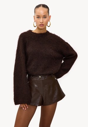Vrouw draagt een donkerbruine, pluizige trui met oversized mouwen en imitatieleren shorts met hoge taille, gecombineerd met grote gouden oorringen.