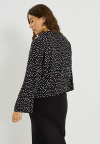 Veste noire courte à pois blancs, dotée de manches longues, d'une texture douce et d'une coupe ample, associée à une jupe noire.