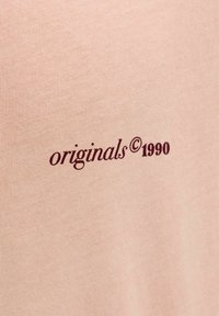 Beežkangas tume punase tekstiga "originals © 1990", mis on trükitud ning kujutab vanamoodsat disainielementi.
