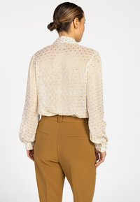 Light-colored, textured blouse with gold dot pattern and long sleeves. Paired with fitted, light brown trousers. Visible shirt pocket.