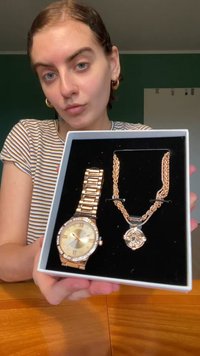 Zelta toņa pulkstenis un atbilstošs kaklarota izstādes kastē. Pulkstenim ir apaļš ciparnīca, romiešu skaitļi un akcenti ar rhinestones. Kaklarota ir ar teksturētu ķēdi.