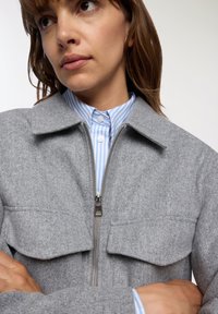 Kobieta z średniej długości brązowymi włosami, ubrana w szary sweter na zamek, nosi niebieską i białą koszulę w paski, z rękami skrzyżowanymi na piersi.