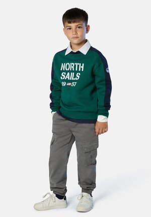 Suéter verde y marino con la gráfica "NORTH SAILS 1957", combinado con pantalones cargo grises y zapatillas blancas. Presenta puños y cinturilla acanalados.