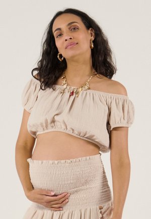 Femme enceinte aux cheveux bouclés portant un haut beige à épaules dénudées et une jupe, tenant doucement son ventre, accessoirisée de boucles d'oreilles et d'un collier en or.